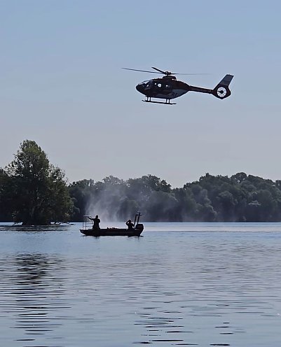 Bild: Ein Rettungshubschrauber im Tiefflug &uuml;ber einem See, auf dem ein Rettungsboot der Feuerwehr mit Besatzung schwimmt. 